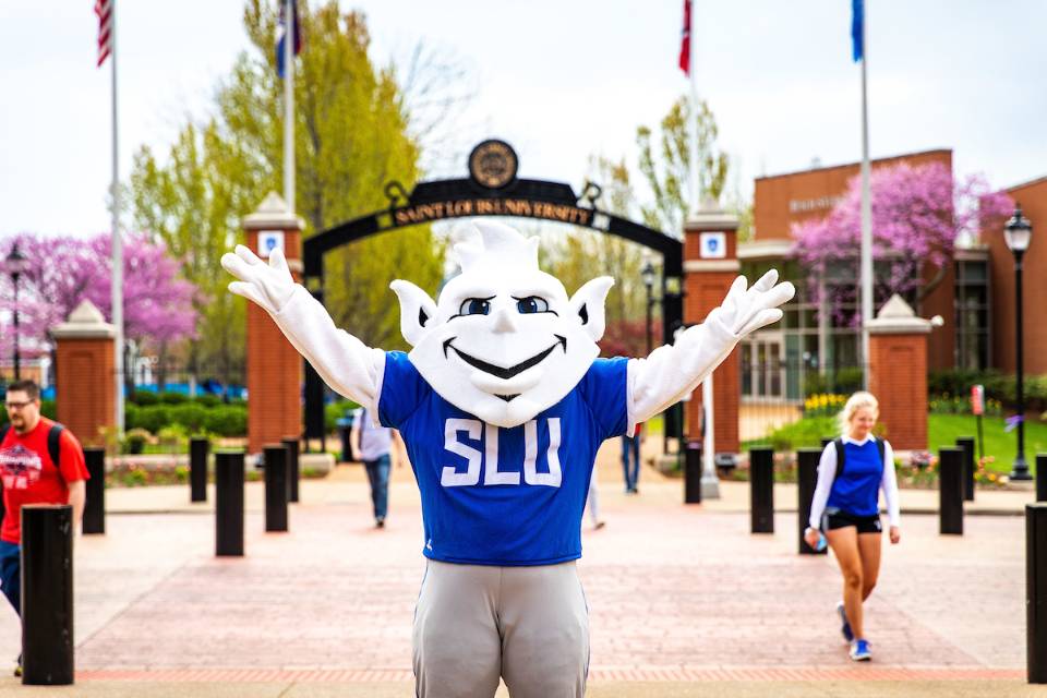 Billiken stands with archway in the background that says Saint Louis University.