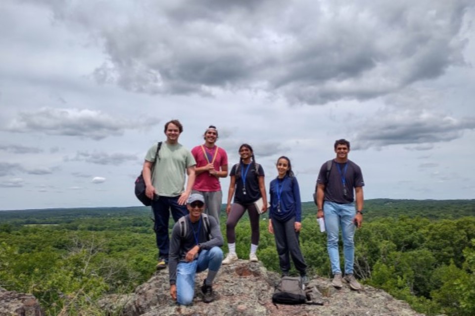 Summer Course Main Photo A group of six students stand and kneel at the top of a rocky bluff overlooking the Ozark forests.