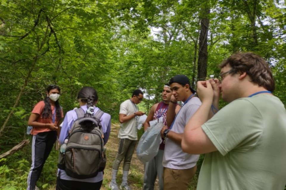 4330-2 A group of students stands in a forest, examining papers and samples.