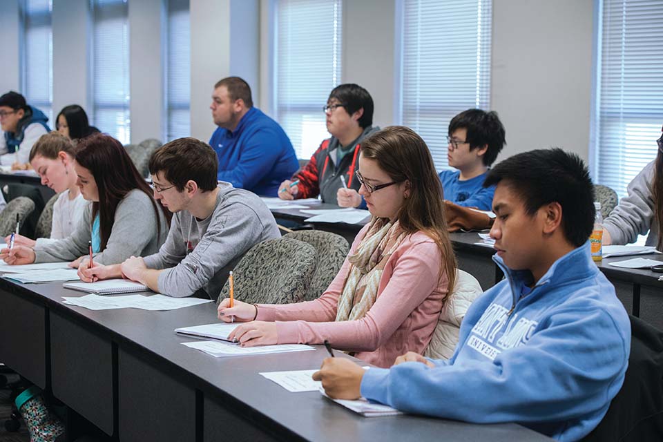 Business School Classroom Students holding pens and pencils sit along a long classroom table, writing on pieces of paper.