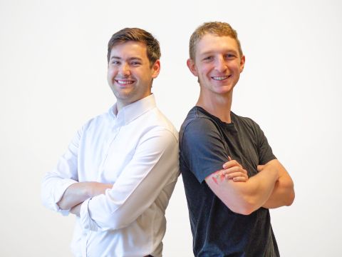 Two men stand back to back smiling with arms folded