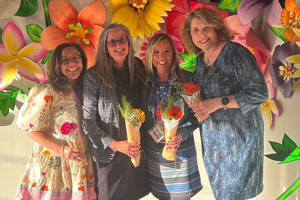 Four women pose in front of a wall covered in flower sculptures. Each one holds a small bouquet.
