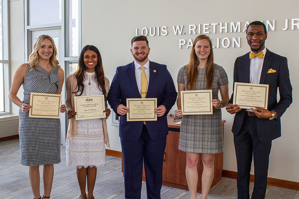 2019 CLEA and David Grant Finalists and Awardees for Excellence in the Legal Clinics Five students wearing formal attire stand in a line. They are holding yellow certifcates for the 2019 CLEA and David Grant finalists awards. They are standing in front of a wall that have silver words spelling out 'Louis W. Riethman Jr'. The students' head are in front of some of the letters.
