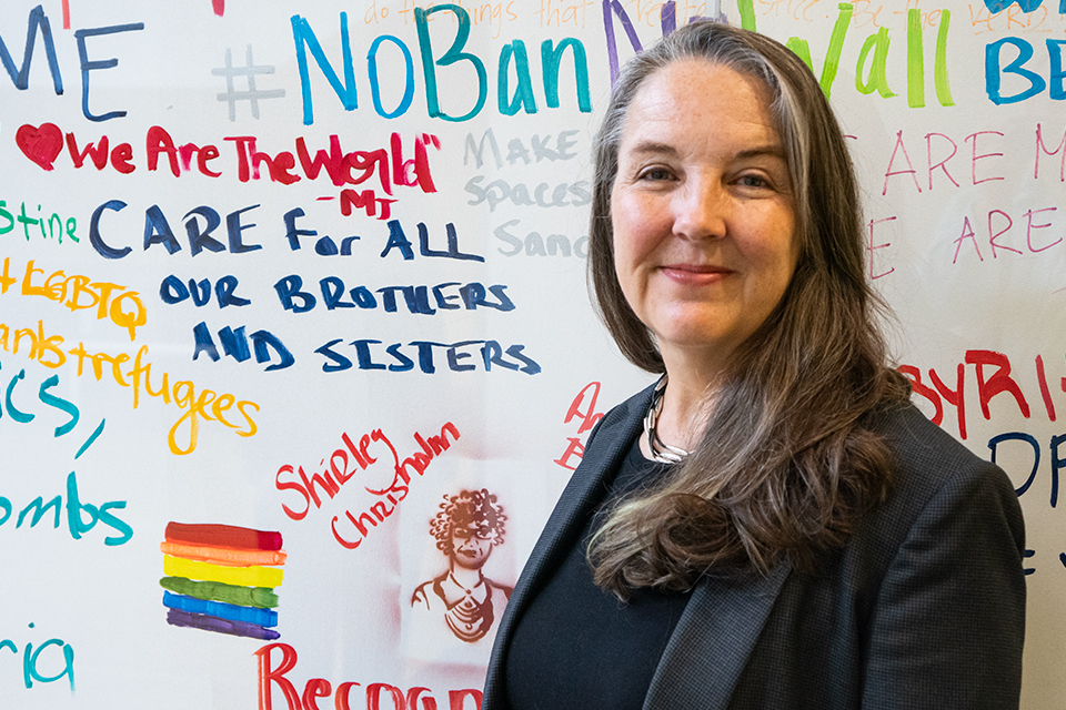 A professor stands in front of a sign with various messages, including Care for all our brothers and sisters, and a drawing of a rainbow flag as well as Shirley Chisholm.