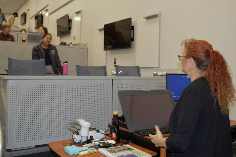 An instructor is seen from behind in a discussion with students sitting in a lecture hall.