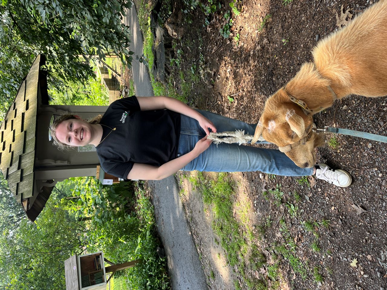 Katherine Tanis stands outside on a wooded walking path with a tan goat