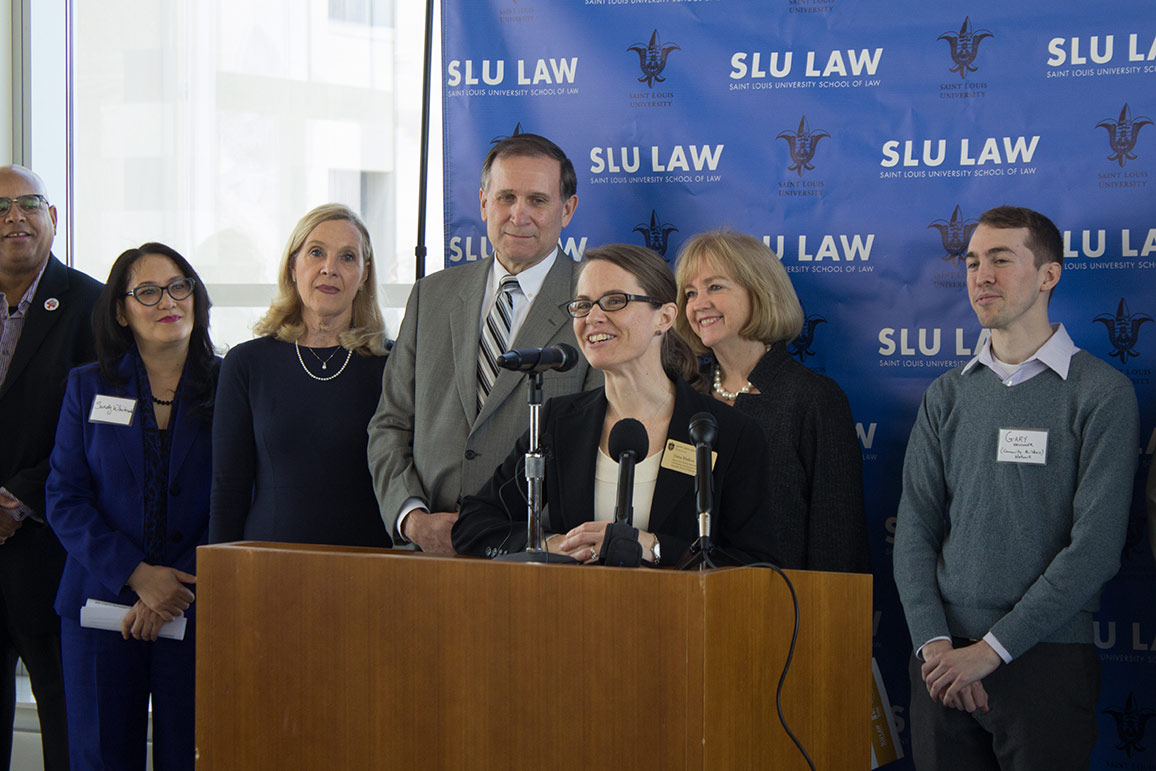 A woman in a suit speaks into a microphone at a lectern while others stand behind her, in front of a SLU Law Banner.