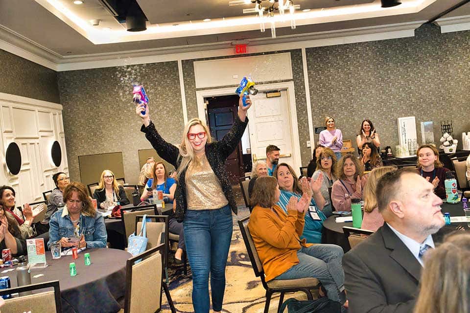 Erika Michalski, a blond woman wearing red framed glasses, holds up two bubble makers while walking past event attendees who are seated at round tables in a large event space.