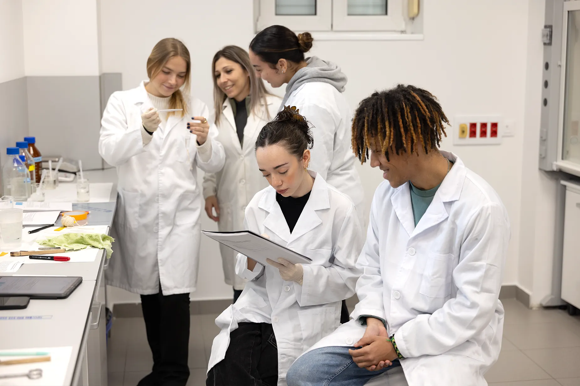A female professor speaks to a small group of students at an outdoor patio. A laptop sits open on the patio table