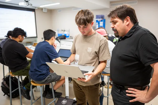 A student, dressed in a graduation gown, shakes hands with a SLU faculty members on stage during commencement. American and Spanish flags are in the background.
