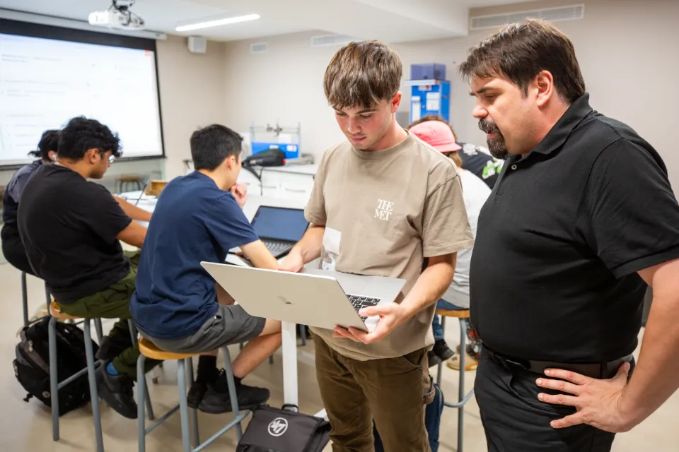 A student and a teacher look at a laptop while other students study seated behind them.
