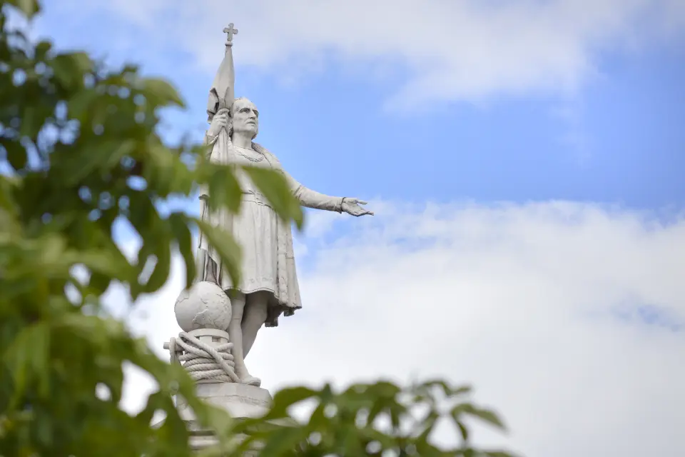 Statue of a person holding a flag, behind the leaves of a tree top.