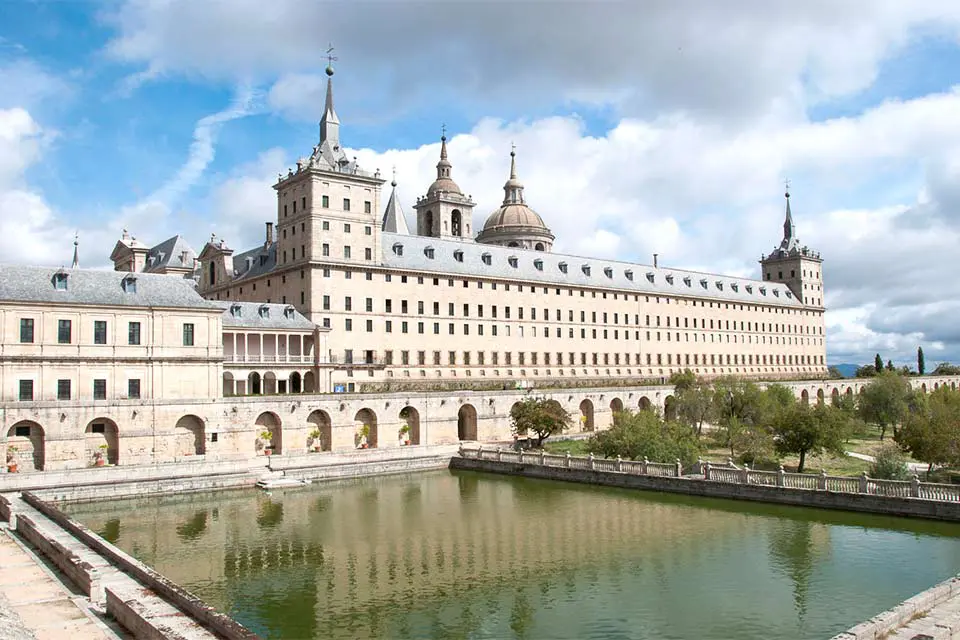Exterior shot of a monastery with a large pond in the patio. The building is made from light bricks and contains multiple arches on the ground floor, with several floors containing rows of windows.