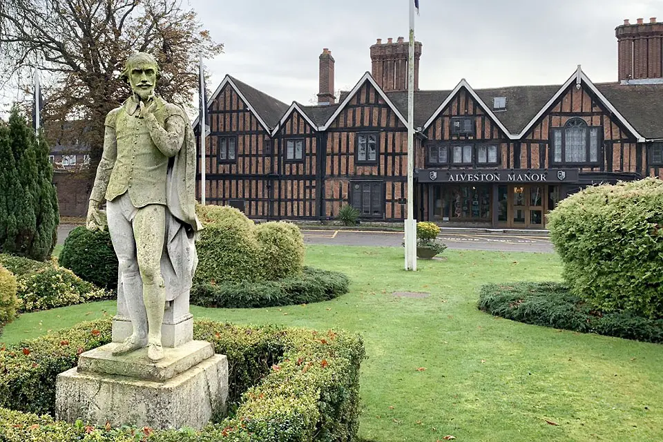 Exterior of a large brick building next to an elaborate garden with a statue of William Shakespeare.