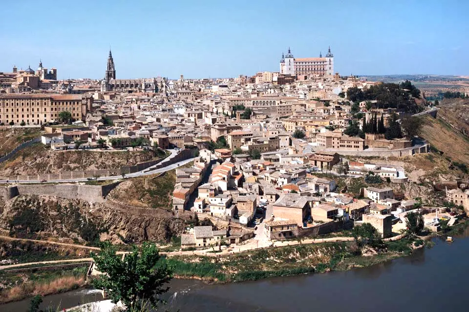Buildings and homes in a city nestle against a hillside next to a river.
