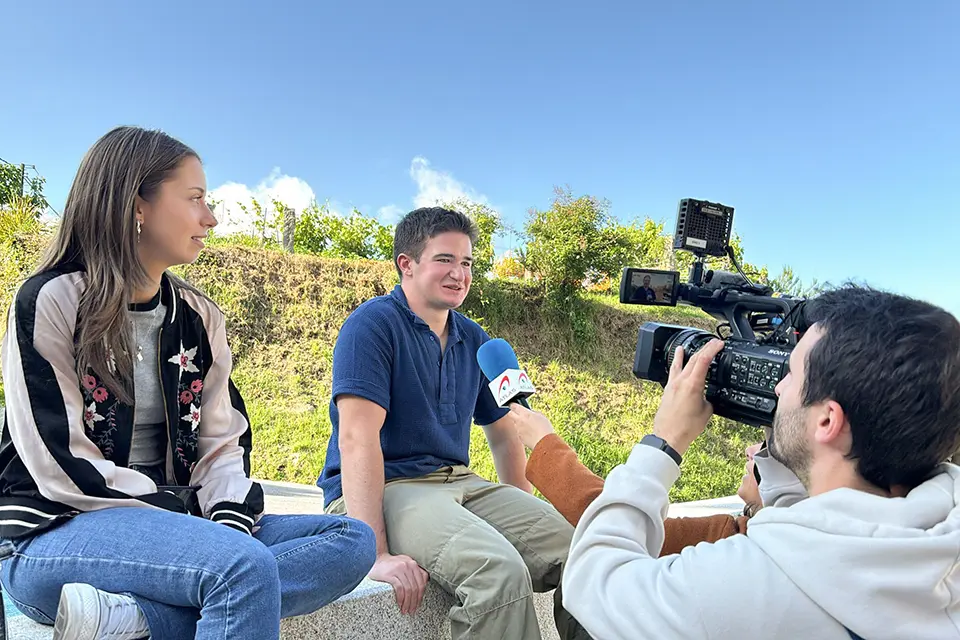 Lara Mitchell Guirao and Alejandro Silvestre Martínez speak to Galician press Two students sitting on a ledge while being interviewed by press that are holding a microphone and camera