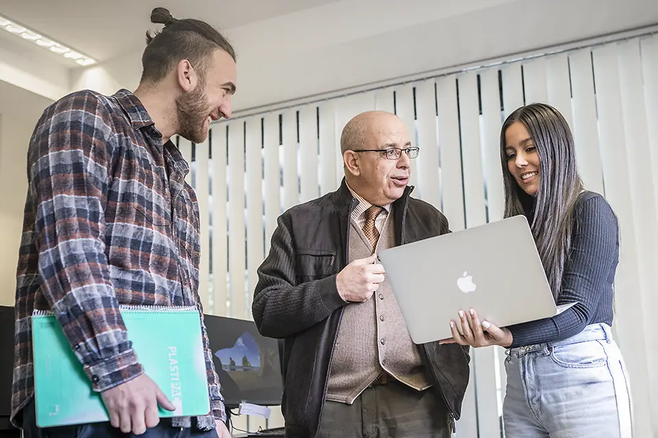 Taieb Gasmi, Ph.D., with students in San Ignacio Hall Computer Lab. Man talking with two students while one of them is holding a laptop.