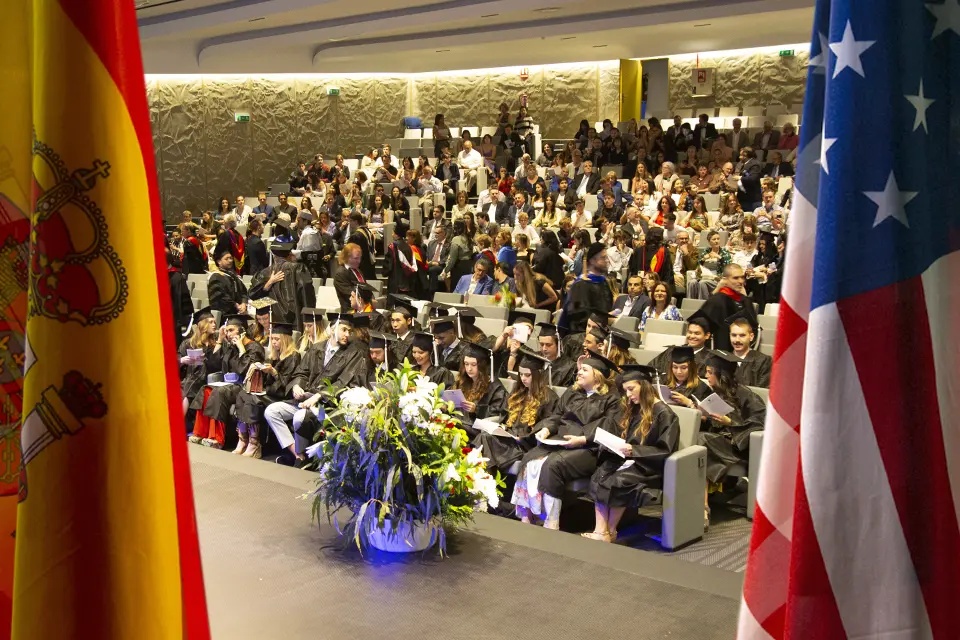 Spanish and U.S. flags frame an image of graduates and sitting in an auditorium wearing caps and gowns and their families.