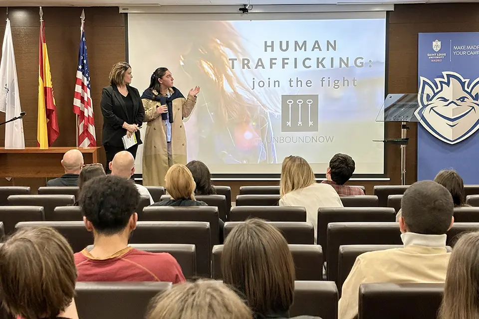 Human Rights Club Takes Action for Women's History Month Student introduces guest speaker on stage in a filled auditorium at the Madrid Campus. A screen with the words "Human Trafficking. Join the Fight. Unbound Now" is visible.