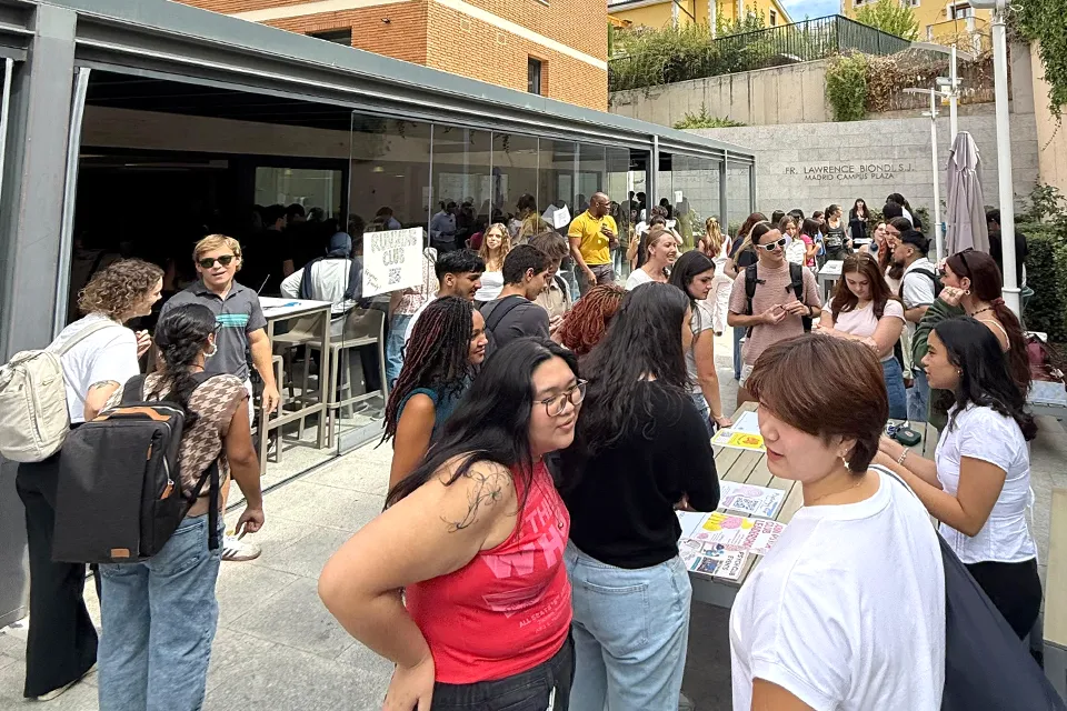 Students gather in the San Ignacio Hall patio. Many students standing around tables in an outdoor patio with a brick wall in the background and doors into a covered patio space.