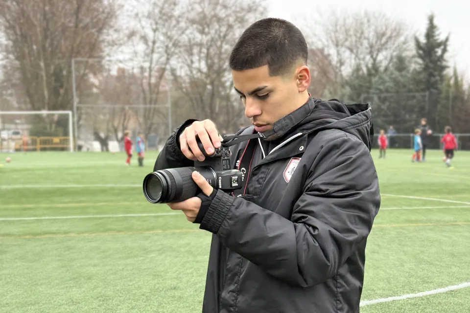 A young man holds a camera on a soccer field with children playing in the background.