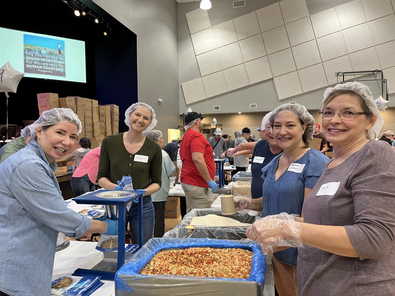 Children 1 Volunteers wearing hairnets and gloves stand around a counter packing items for meals.
