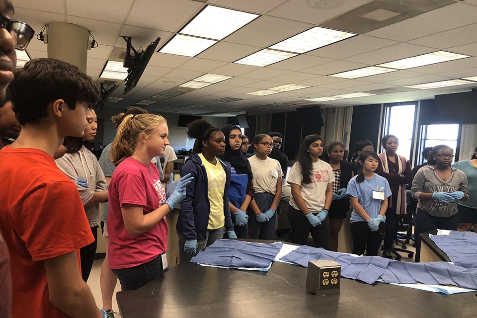 Students participating in pathology hands-on lab High school students wearing latex gloves stand around a lab table.