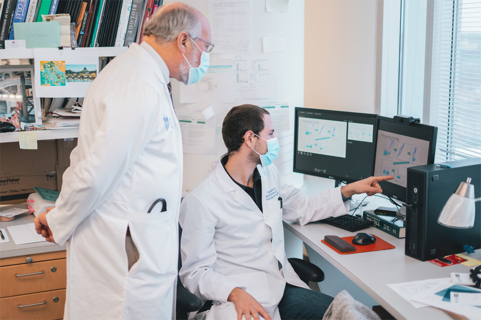 Professor Daniel Hoft supervising student on a presentation creation A student sits and points at a computer screen while a professor stands behind him supervising.