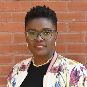 A shoulder-length portrait of a woman with textured black hair, closely cropped on the sides, stands in front of a brick wall. She wears glasses with a blue tortoise-shell pattern, gold earrings, a gold chainlink necklace, and a black top with a floral-print jacket. Her smiles slightly gazes with a pleasant expression.