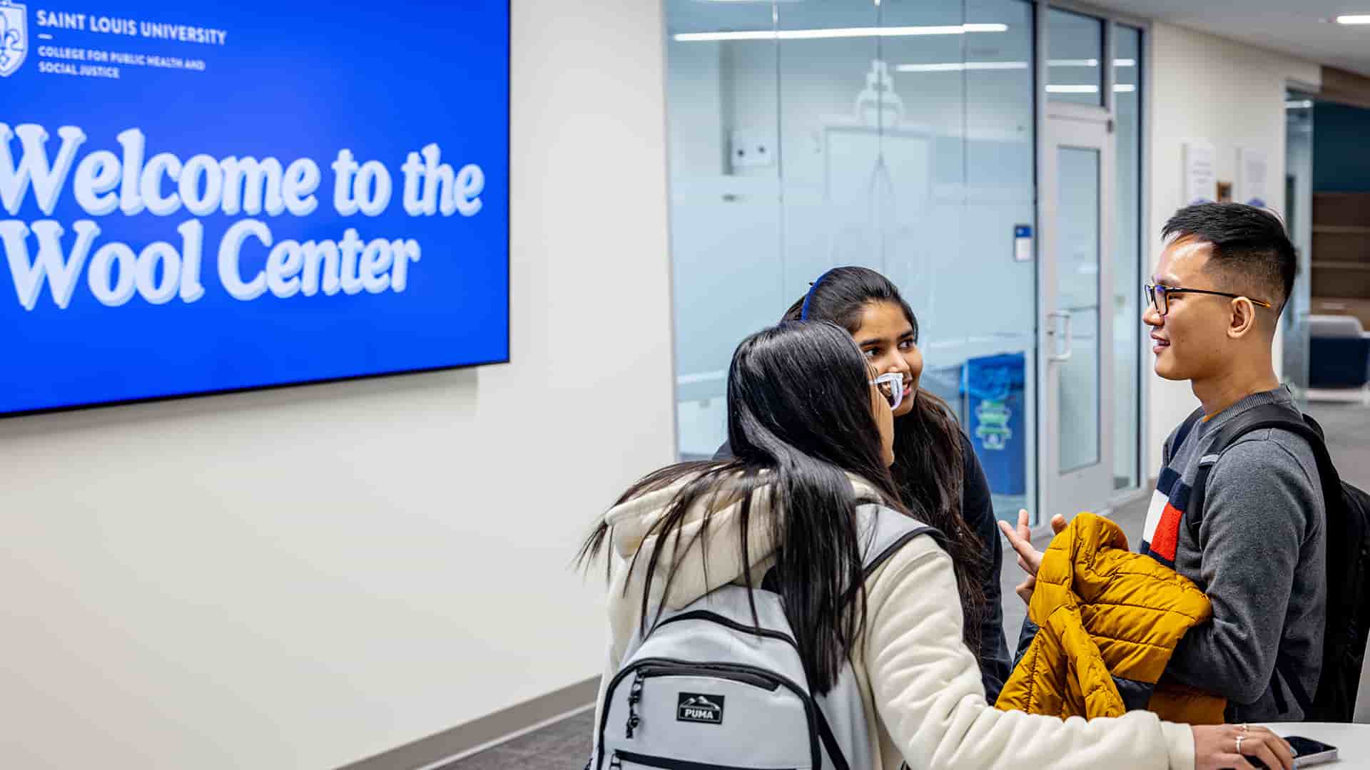 Three students gather outside of a classroom along a hallway of glass walls.