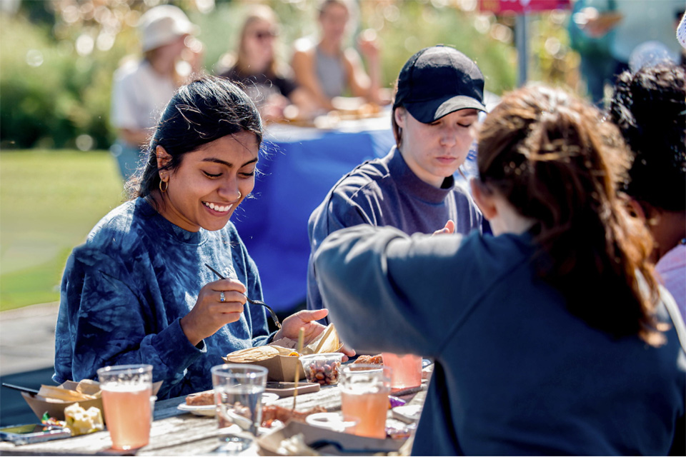 SLU students enjoy a meal outside. SLU students sit at a picnic table filled with cups and cardboard trays filled with food on a sunny day.