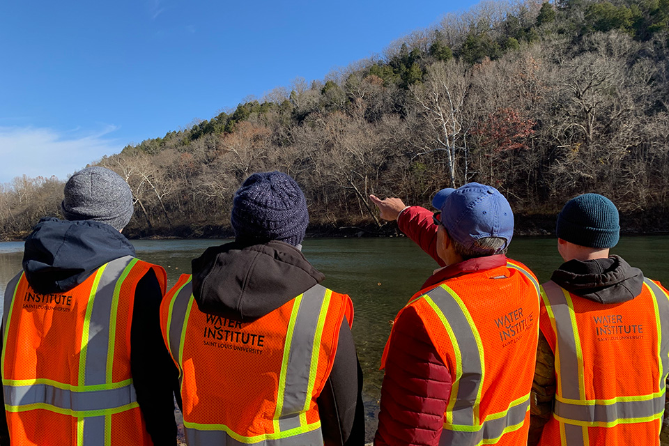 Four team members are shown from behind, wearing reflective vests that read Water Institute. One person points toward the distance as they look across the river at a shoreline of leafless trees.